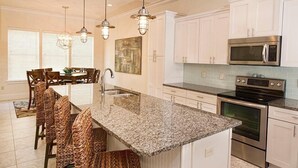 A kitchen with white cabinets, a granite countertop, a stainless steel microwave, and an oven. There are wicker chairs around the island and pendant lights above. - LA TERRAZZA 948A 4 Bedroom Holiday Home by Five Star Properties (Miramar Beach)
