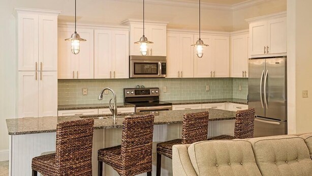 A kitchen with white cabinets, a stainless steel refrigerator, a microwave, and a built-in oven. There are three wicker chairs and a bar stool around the island. - LA TERRAZZA 948A 4 Bedroom Holiday Home by Five Star Properties (Miramar Beach)