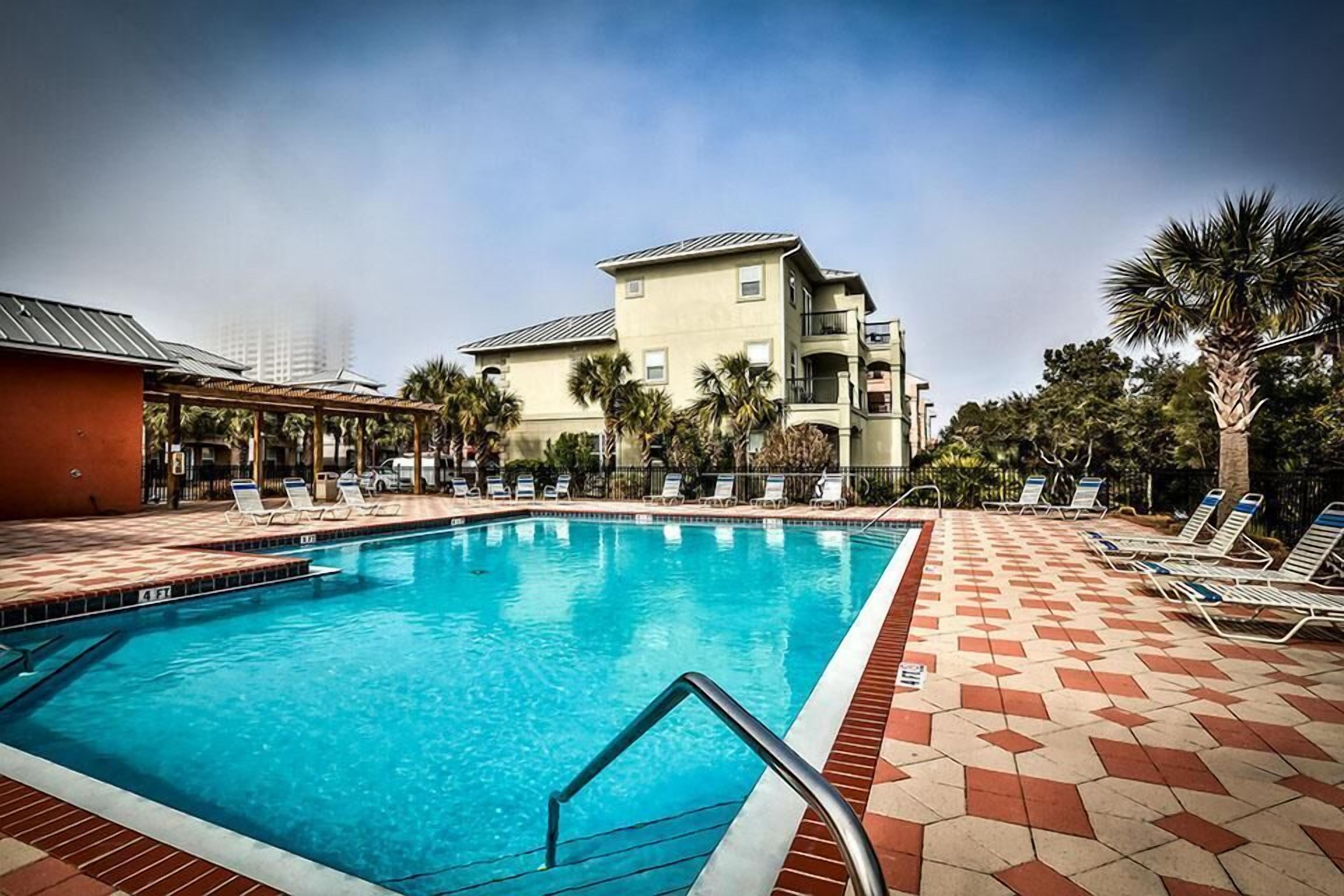 A swimming pool with lounge chairs, a covered area, and a multi-story building in the background.