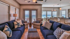 A spacious living room with a ceiling fan, a dining area with chairs, and a view of the ocean through large windows.