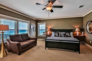 A bedroom with a bed, a ceiling fan, a nightstand, a lamp, and a view of the ocean through large windows.