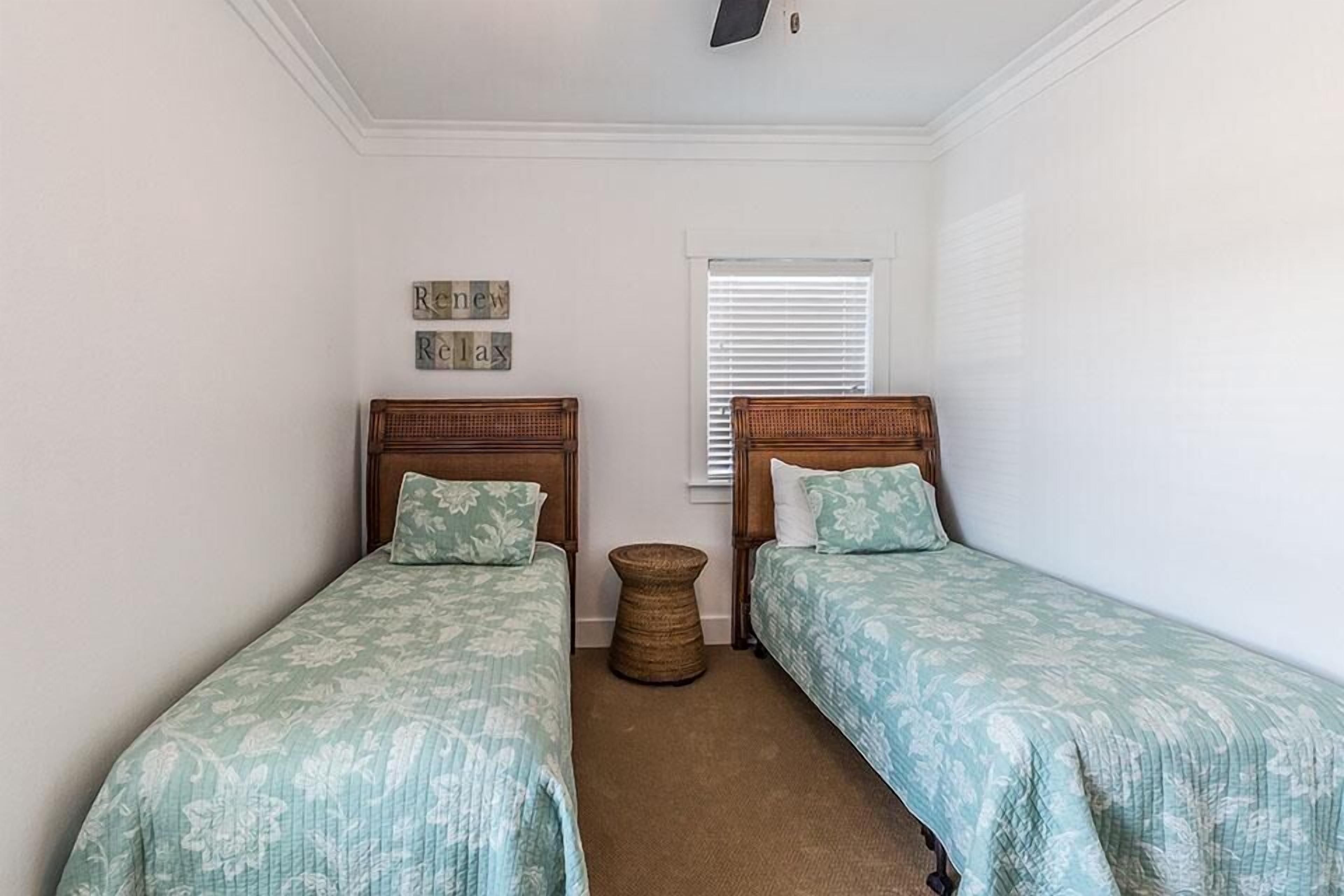 Un dormitorio con dos camas individuales, un taburete redondo de mimbre y una ventana con persianas.