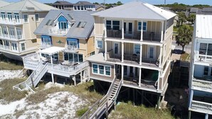A multi-story beachfront house with balconies, a staircase leading to the beach, and a sandy foreground.