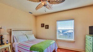 Un dormitorio con cama, ventilador de techo, ventana con persianas, mesita de noche y cómoda.