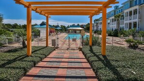 Um caminho leva a uma área de piscina sob uma pérgula de madeira, cercada por vegetação e um cercado.