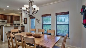 Uma área de refeições com mesa e cadeiras de madeira, um candelabro e uma cozinha ao fundo.