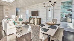 A spacious living room with a high ceiling, a dining area with a wooden table and chairs, a white sofa set, a chandelier, and a large abstract painting on the wall. - Looking Glass 5 Bedroom Holiday Home by Five Star Properties (Santa Rosa Beach)