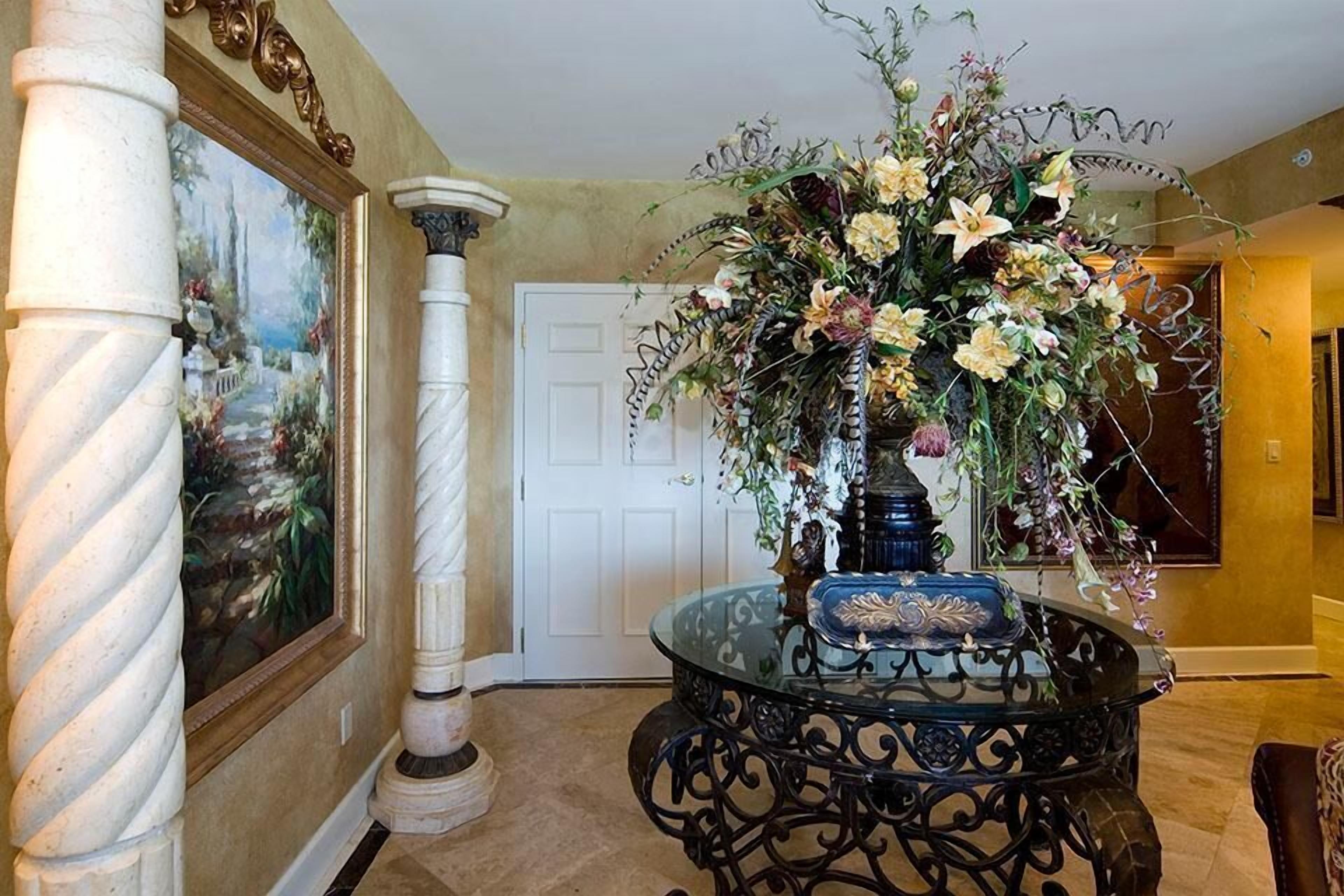 A lobby with a round glass table, a large floral arrangement, a painting, and a white door.
