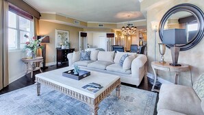 A spacious living room with a large sofa, a coffee table, and a chandelier. There is a dining area in the background with a chandelier and a large mirror. - MEDITERRANEA 401A 3 Bedroom Holiday Home by Five Star Properties (Miramar Beach)