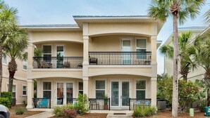 Um edifício residencial de dois andares com telhado plano, varandas com guarda-corpos pretos e um pequeno jardim frontal com plantas e um banco.