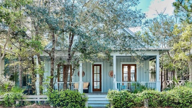 Une maison bleu clair avec un porche, entourée d’arbres et d’une clôture blanche en bois.