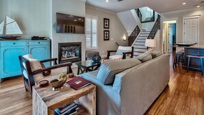 A spacious living room with a sofa, wooden coffee table, and a fireplace. A staircase leads to an upper level, and a kitchen area is visible in the background. - Ocean Escape 4 Bedroom Holiday Home by Five Star Properties (Destin)