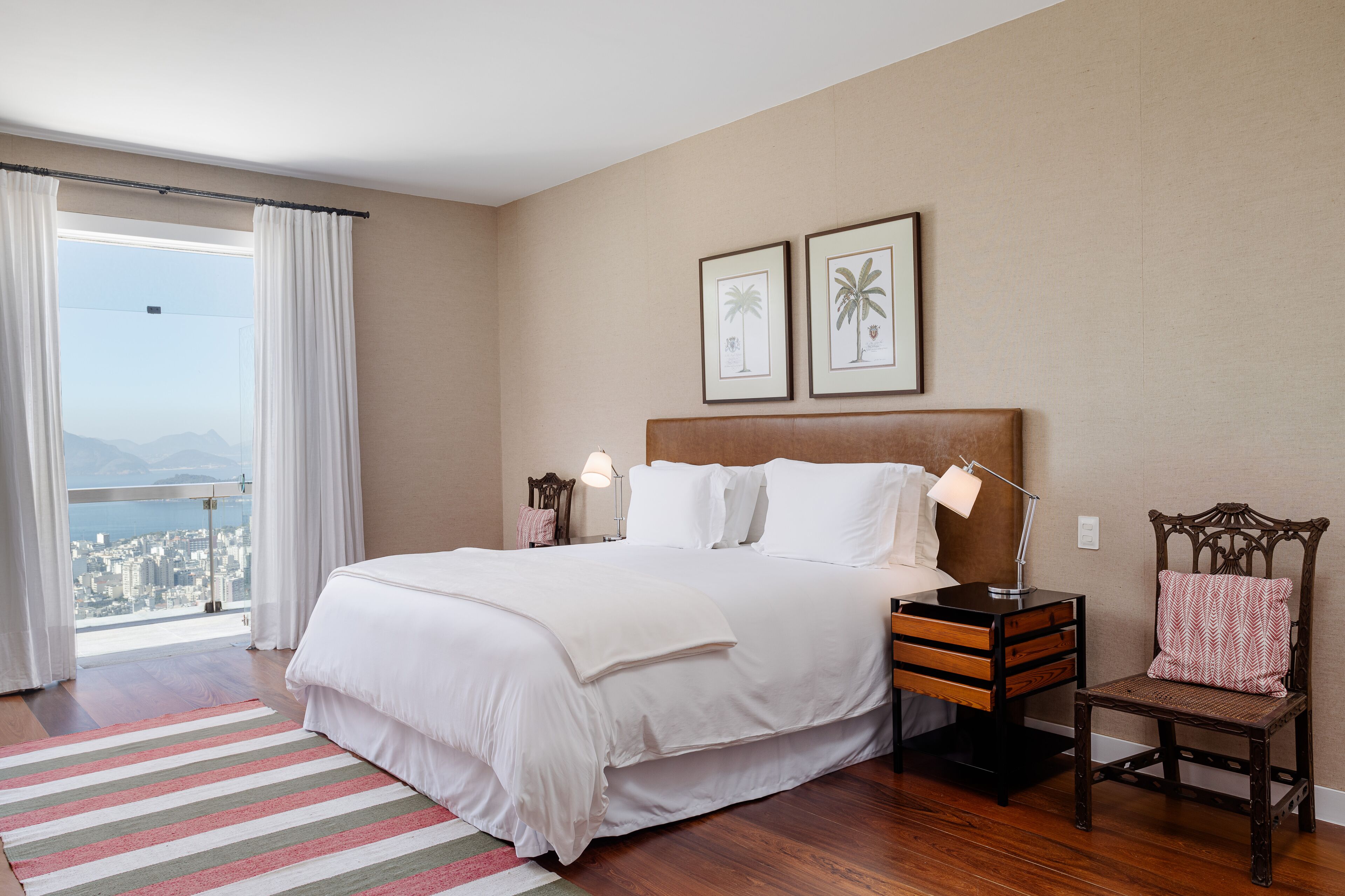 A hotel room with a large bed, a wooden nightstand, a chair, and a view of the city through a balcony.