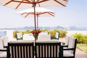 Eine Terrasse mit Tisch und Stühlen unter einem großen Sonnenschirm, mit Blick auf ein Gewässer und Berge.