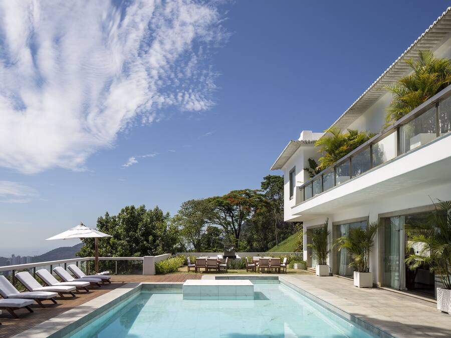 Un área de piscina moderna al aire libre con sillones reclinables y plantas en macetas, rodeada por un edificio blanco con amplias ventanas y un balcón con vistas a un paisaje pintoresco.