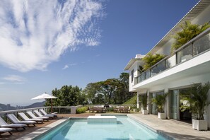 Ein moderner Poolbereich im Freien mit Liegestühlen und Topfpflanzen, umgeben von einem weißen Gebäude mit großen Fenstern und einem Balkon mit Blick auf eine reizvolle Landschaft.