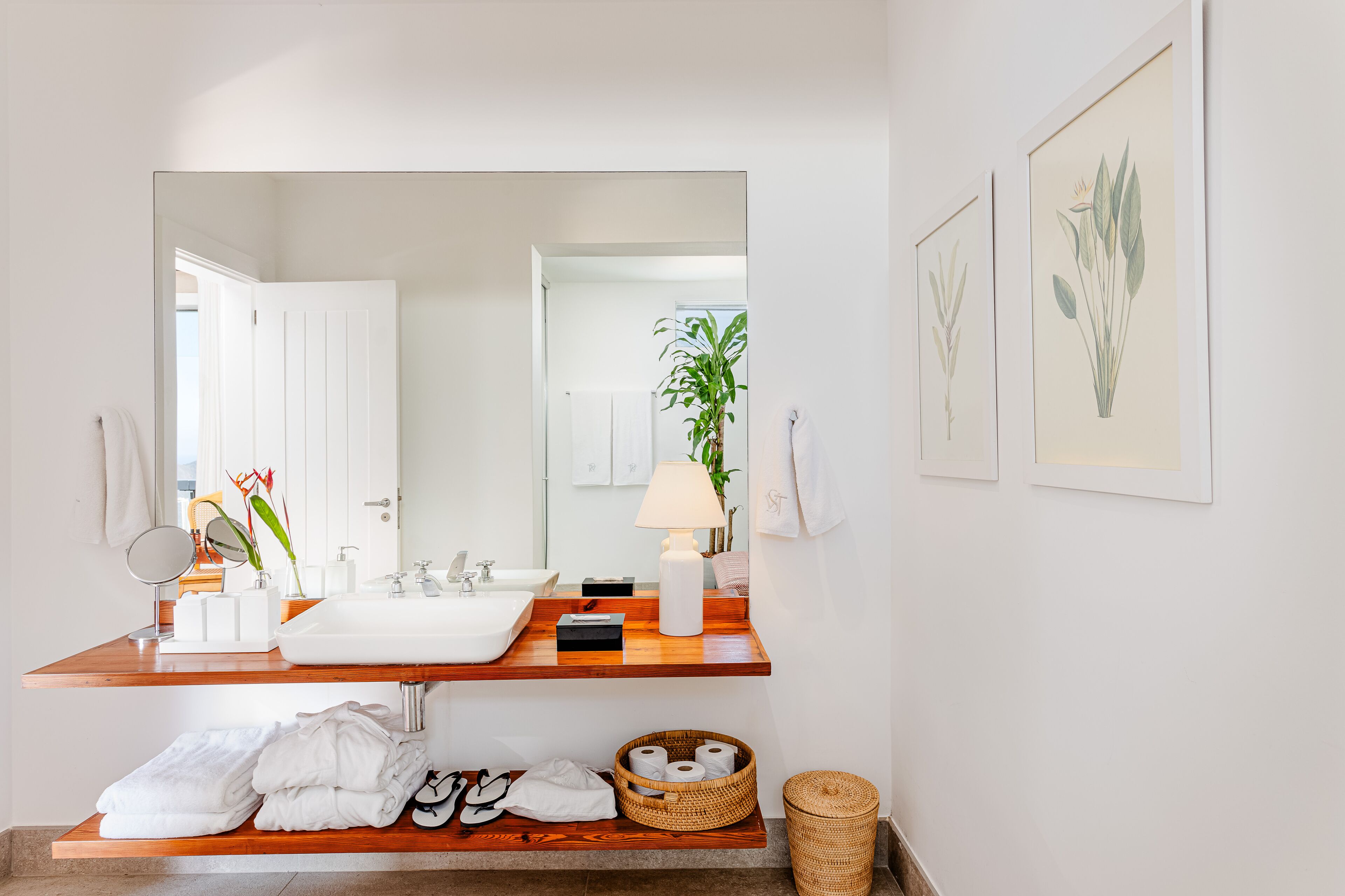 Un baño con tocador de madera, lavabo blanco y espejo. Hay toallas, un cesto de artículos de aseo y una planta.