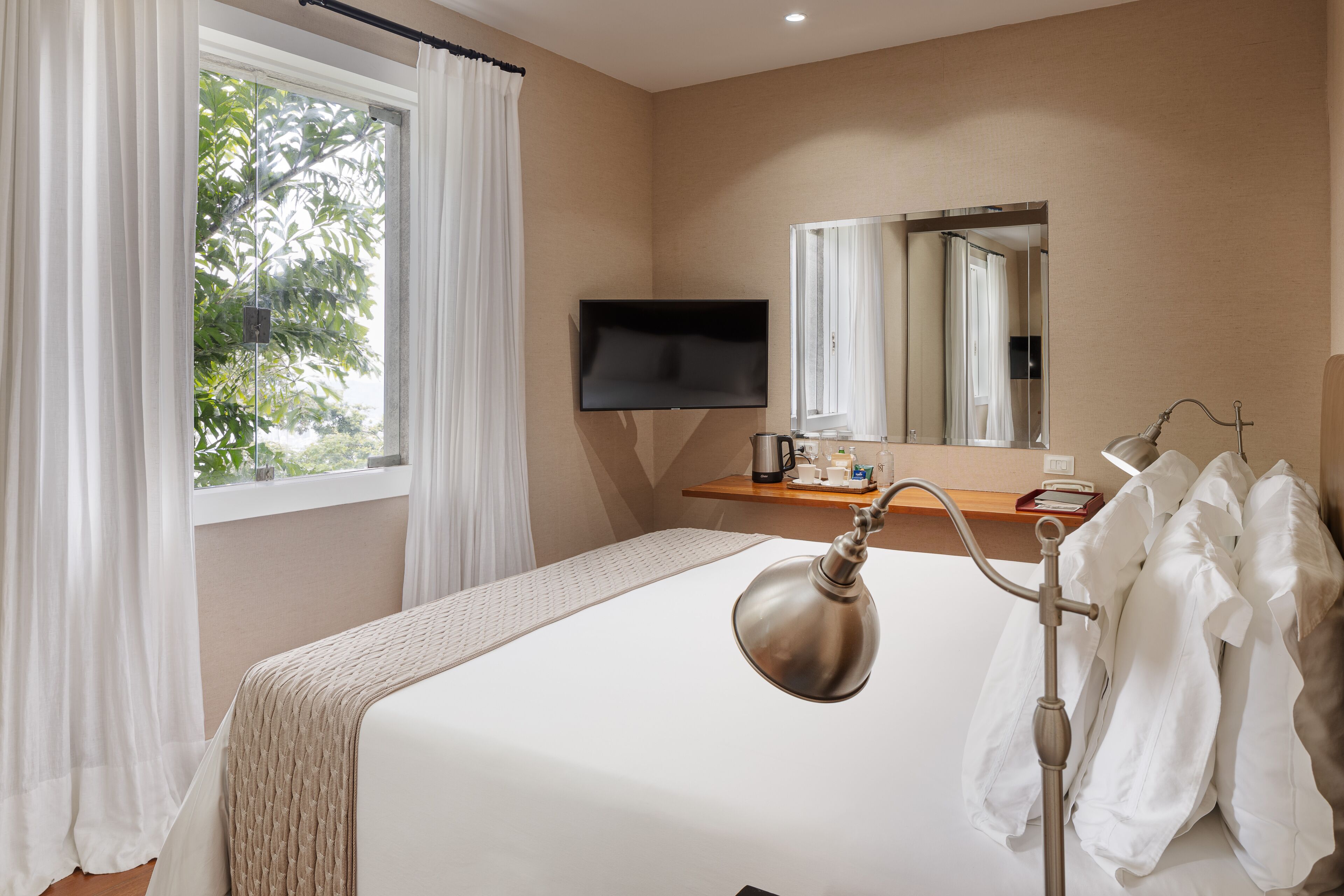 Ein Hotelzimmer mit Bett, Fernseher, Nachttisch mit Lampe, Spiegel und einem Fenster mit Vorhängen.