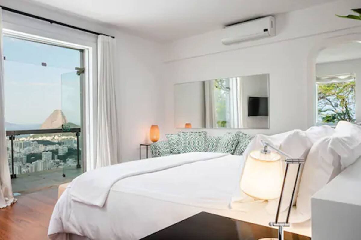 Una habitación de hotel moderna con una cama grande, una mesita de noche con lámpara, televisión y vistas a la ciudad desde un balcón.