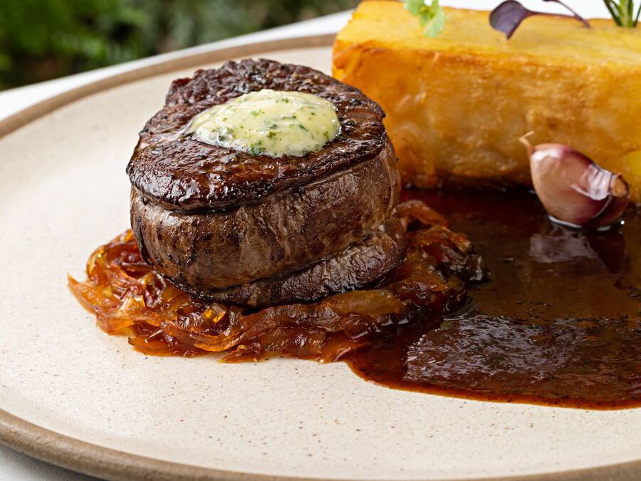 Un bistec con un poco de mantequilla encima, acompañado de cebollas caramelizadas y una guarnición de papas fritas.