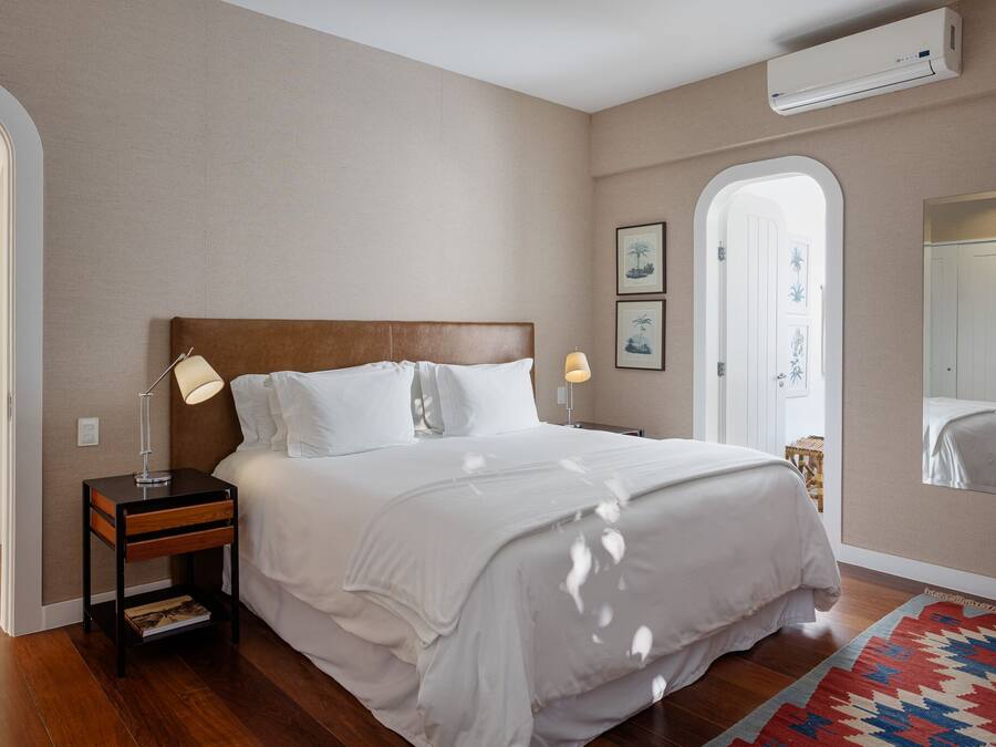 A bedroom with a wooden headboard, white bedding, a nightstand with a lamp, and a patterned rug.