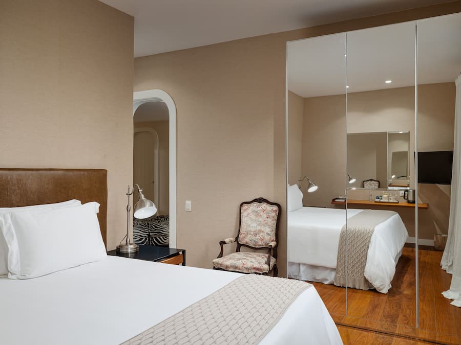 A hotel room with a large bed, a chair, a desk with a lamp, and a mirror.