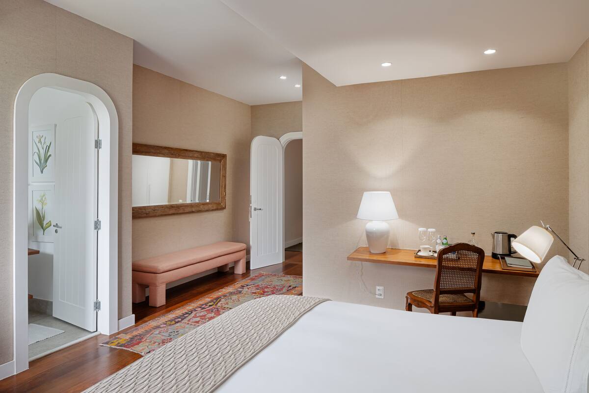Habitación de hotel con cama, escritorio con lámpara, silla, banco y espejo.