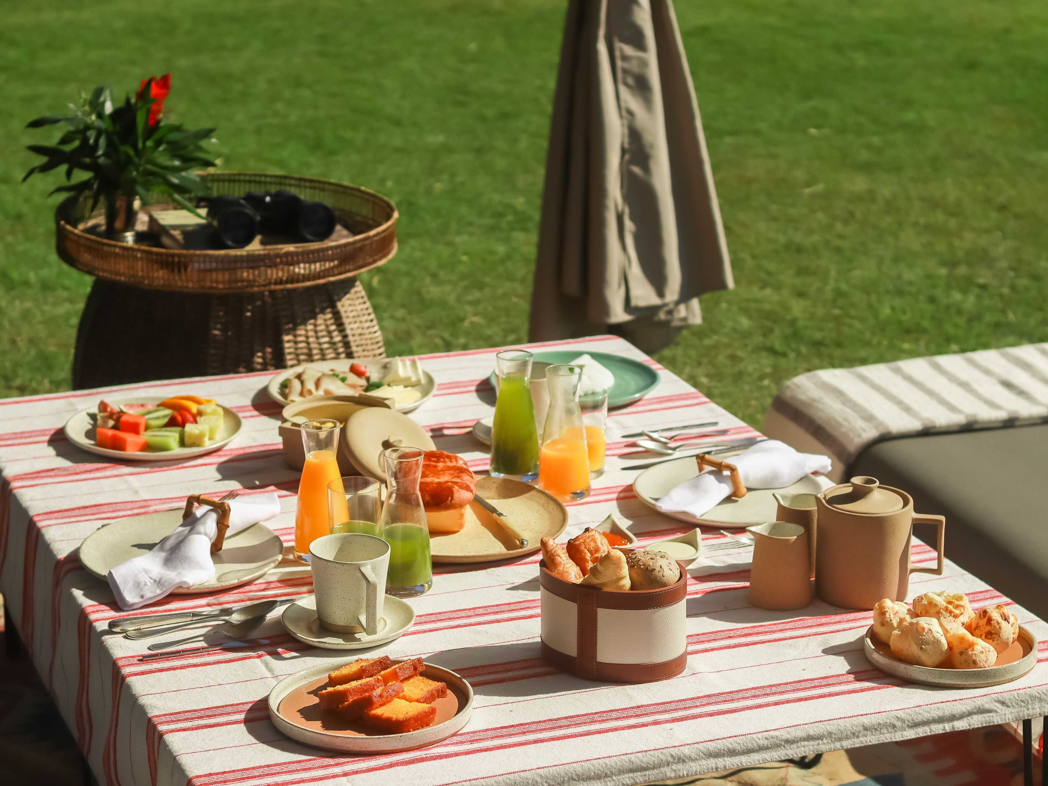 A picnic setup with a table covered by a striped tablecloth, featuring plates of food, drinks, and a basket of bread.