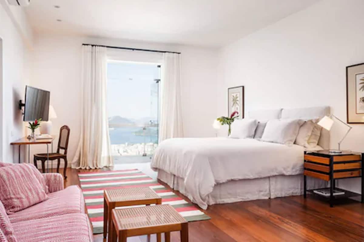 Un dormitorio con cama, escritorio, silla, sofá, ventana con cortinas y vista al mar.