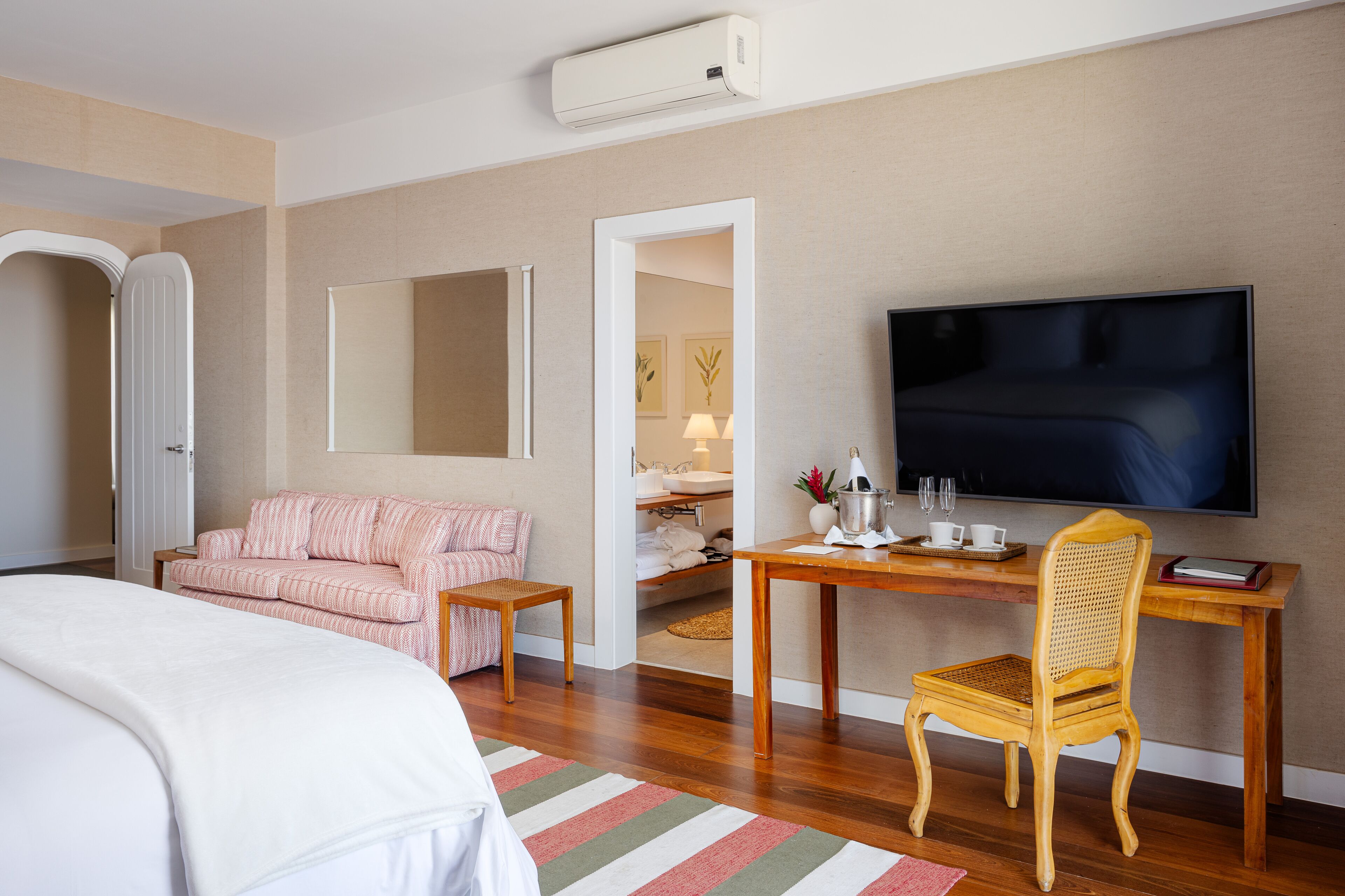 A hotel room with a bed, a pink striped sofa, a wooden desk with a chair, a flat-screen TV, and a bathroom visible through an open door.