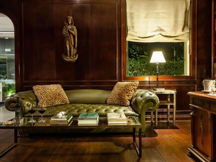 Uma sala de estar aconchegante com um sofá de couro verde, almofadas com estampa de leopardo, uma mesa de centro com livros e copos, uma estátua na parede e uma janela com uma luminária.