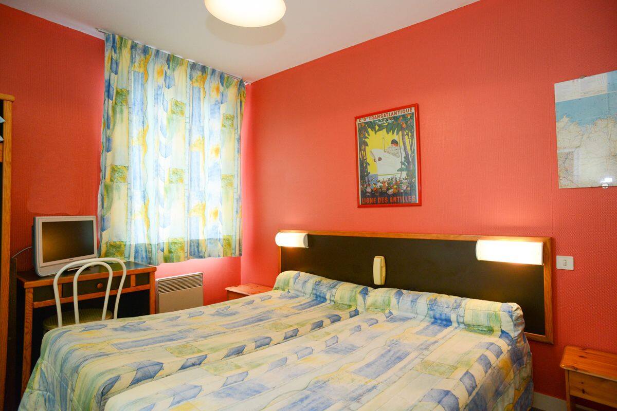Ein Schlafzimmer mit einem Bett, einem Schreibtisch mit Computer, einem Stuhl, einem Fenster mit Vorhängen und einer Wand mit einer Karte und einem Poster.