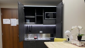 A compact kitchen with a microwave, sink, and cabinets, a dining table with yellow chairs, and a potted orchid. - Hotel Rinaldo Apartments (Borgomanero)