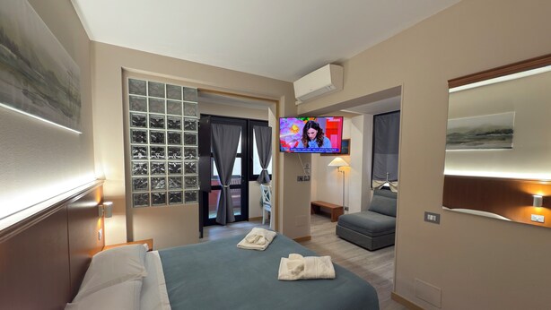 A hotel room with a bed, a television, a seating area, and a view of the outside. - Hotel Rinaldo Apartments (Borgomanero)
