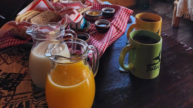 A breakfast table with bread, butter, jam, and coffee mugs in a wooden cabin with a view of the sky and mountains. - Cabañas Alechen (El Calafate)