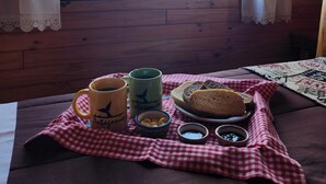 A cozy cabin room with a large window, a checkered tablecloth, a plate of bread, two mugs, and a bowl of pasta. - Cabañas Alechen (El Calafate)