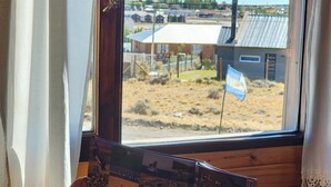 A person reading a book by a window with a view of a rural landscape and a flag. - Cabañas Alechen (El Calafate)