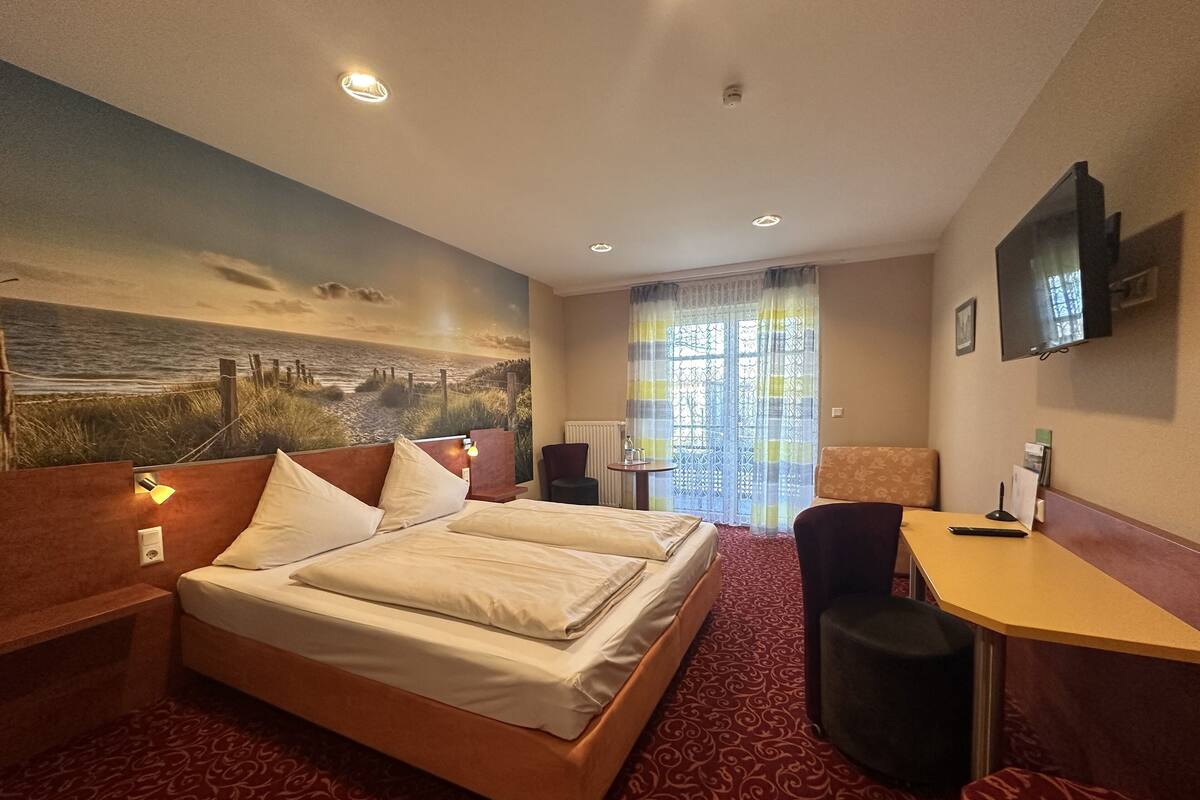 A hotel room with a large bed, a desk, a chair, a TV mounted on the wall, and a painting of a beach scene on the wall.