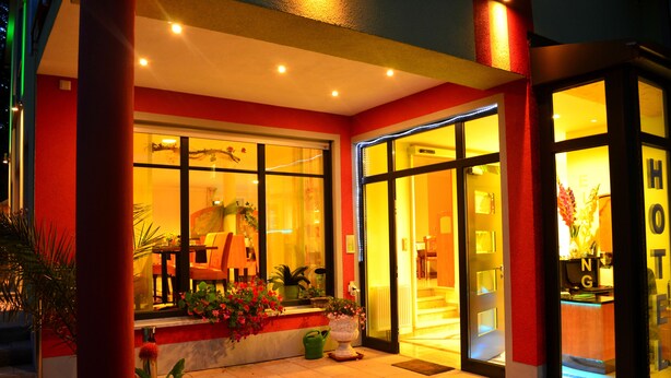 A hotel entrance with a red facade, large glass windows, and potted plants. - Rothsee Hotel (Allersberg)