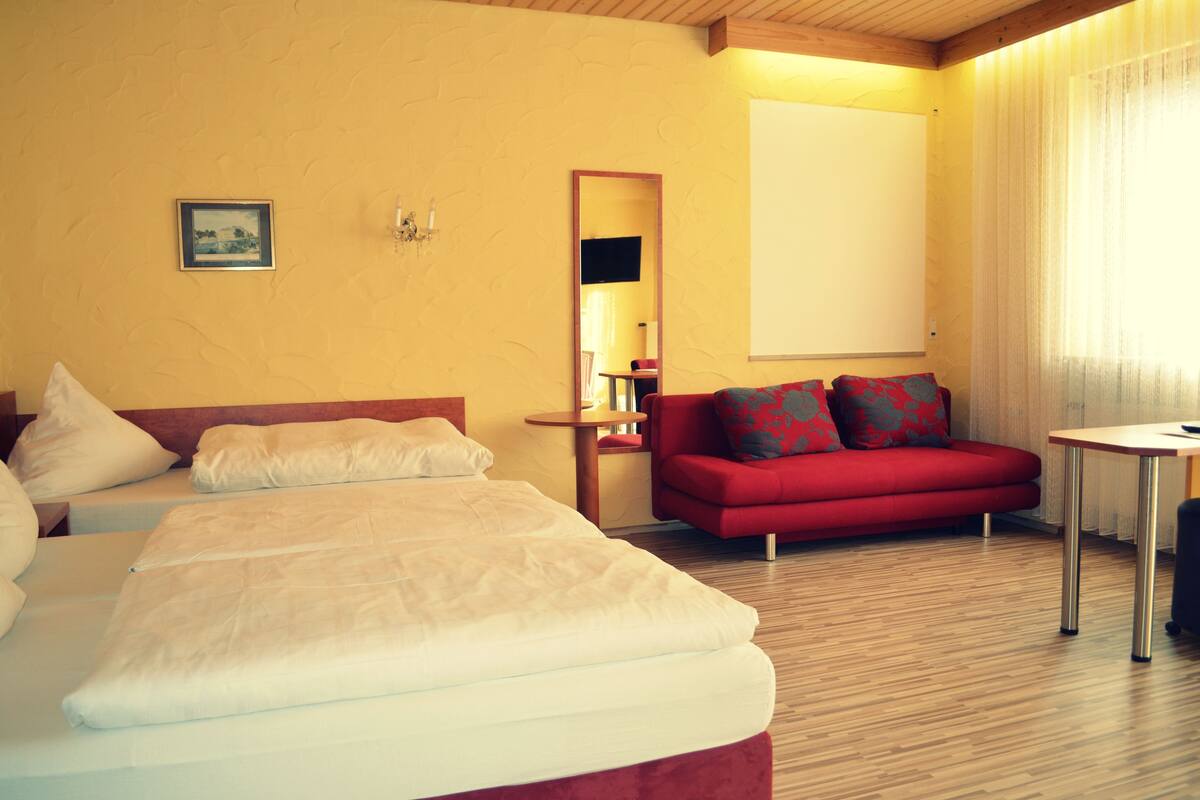 A hotel room with a bed, a red sofa, a small table, and a mirror.