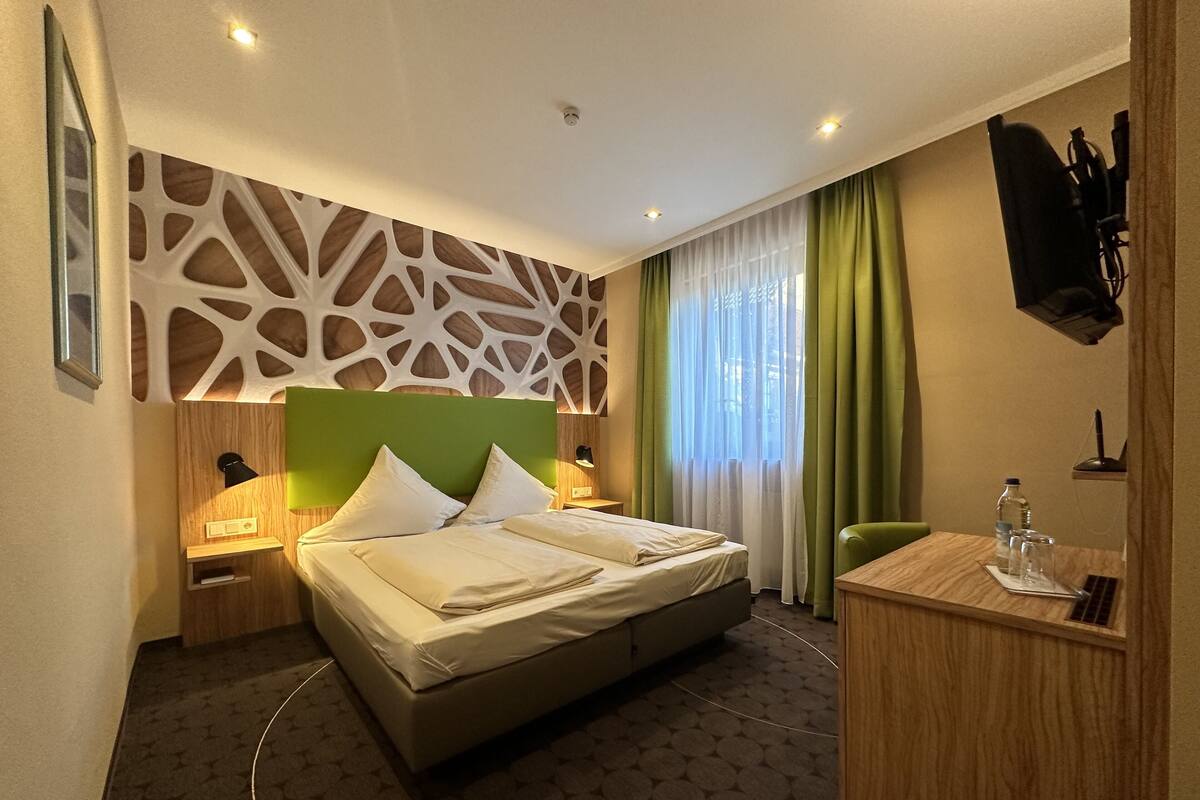 A hotel room with a bed, bedside tables, a desk, a chair, a TV, and a window with curtains.