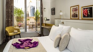 Habitación de hotel con cama, una silla, una mesa y un balcón con vistas a edificios y árboles.