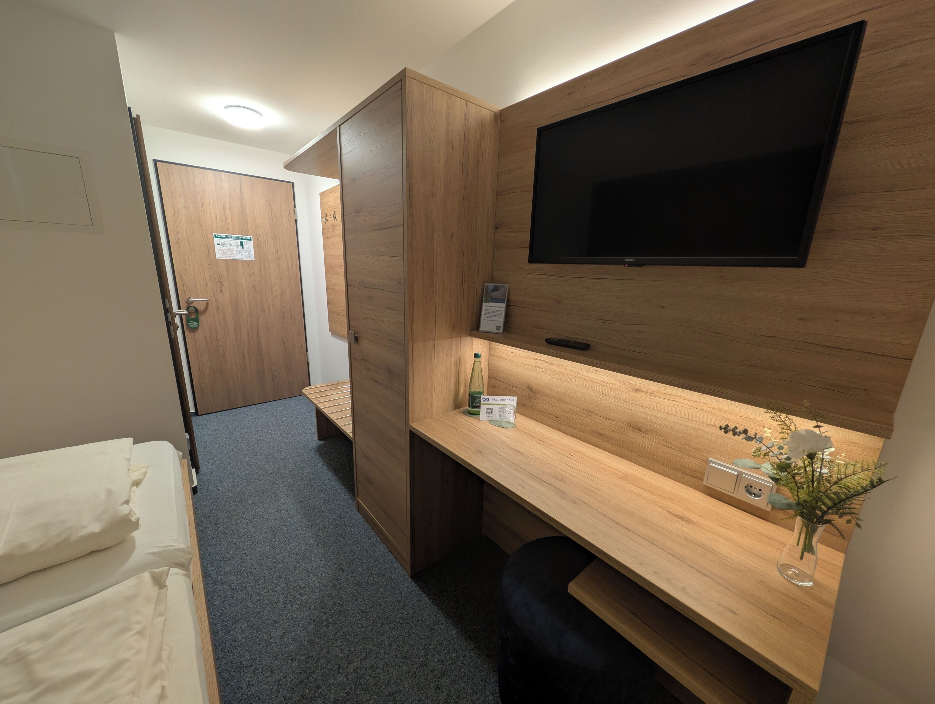 Una habitación de hotel con una cama, un escritorio con televisor, un armario de madera y un pequeño banco.