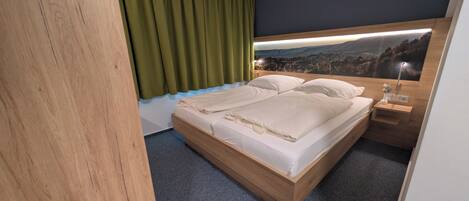 A double bed with a wooden frame, a nightstand with a vase, and a view of a landscape through a window with curtains.