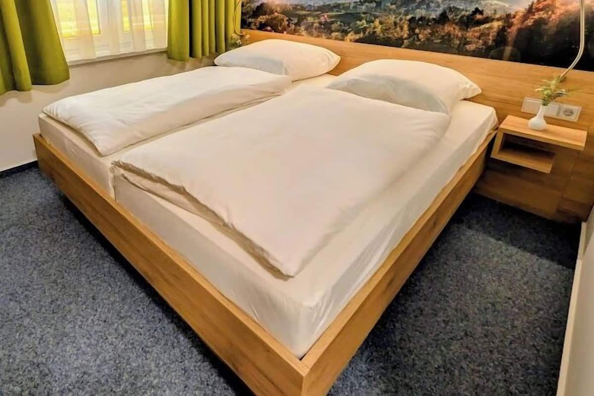 Una cama doble con estructura de madera, una mesita de noche con lámpara, y vista a un paisaje montañoso a través de una ventana con cortinas verdes.