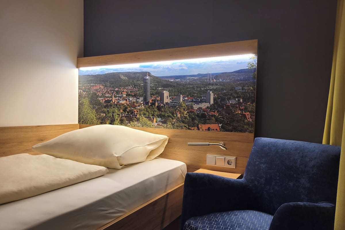 Habitación de hotel con cama, una silla y un televisor de pared que muestra una vista panorámica de la ciudad.