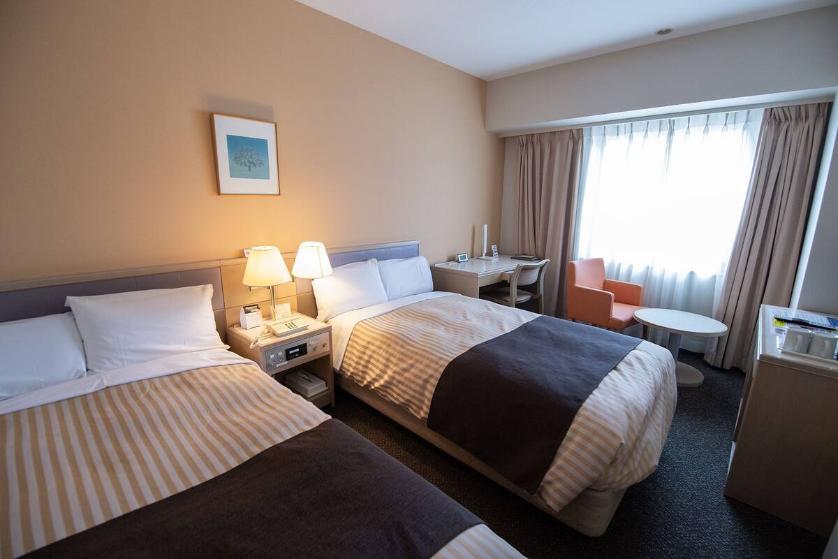 A hotel room with two beds, a desk, a chair, a small table, and a minibar.