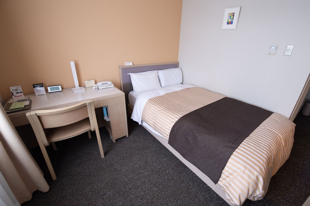 A hotel room with a bed, a desk, a chair, a clock, and a thermostat.