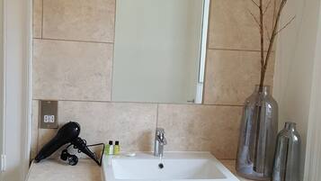 A bathroom with a white sink, a mirror, and a hairdryer on the counter. There are three bottles of shampoo and conditioner on the sink.