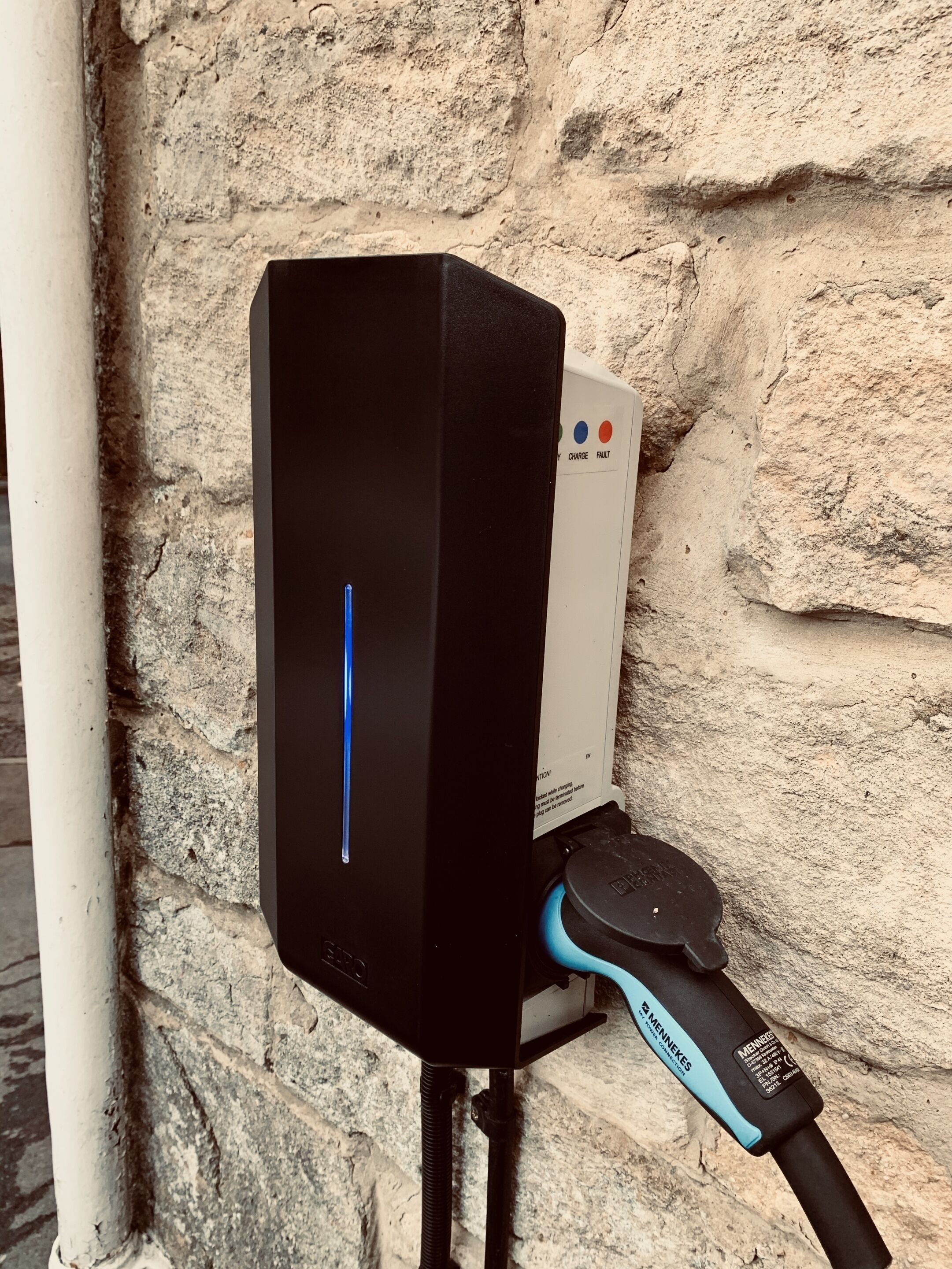 Estación de carga para vehículos eléctricos negros, con indicador luminoso azul, conectada a una unidad de carga blanca con un enchufe.